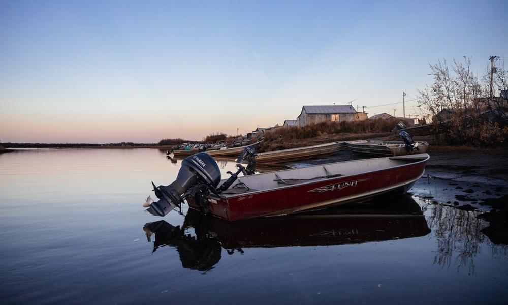 A boat with its motor raised is moored on the coast at sunset with small buildings in the background.